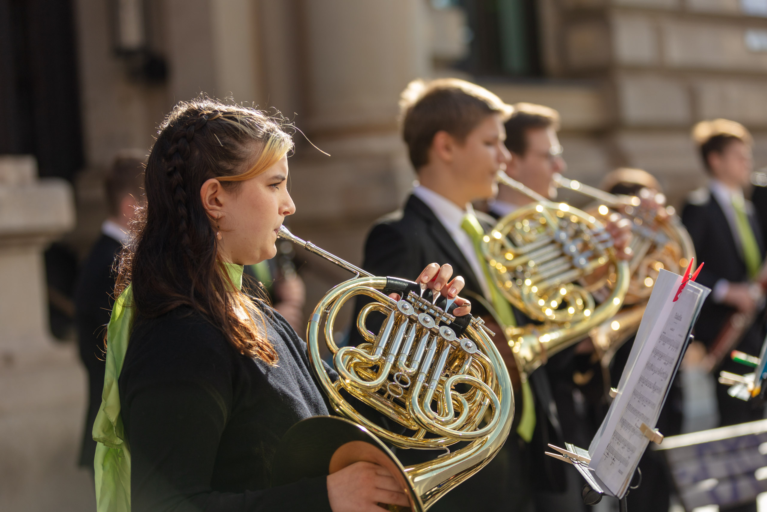 Mitglieder des Jugendsinfonieorchster, die draußen vor der Musikschule Waldhorn spielen, im Vordergrund eine junge Frau mit grünem Tuch, die Horn spielt
